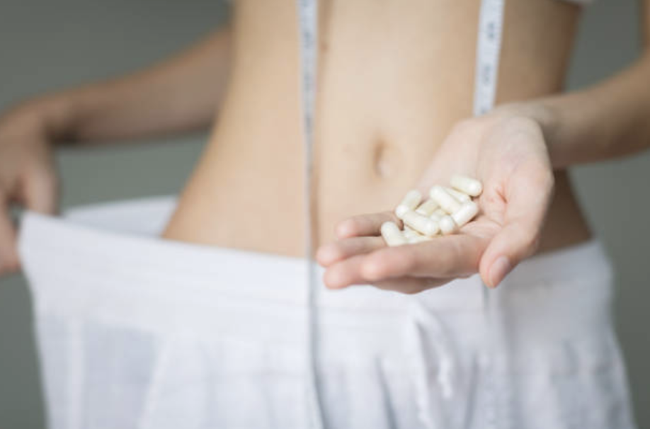 Person holding weight loss pills with measuring tape around slim waist